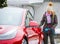 Stylish girl stands near her red electric car and waits for the car to charge. Connecting the plug of the electric