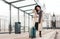 Stylish girl with a suitcase stands at a city stop awaiting a tram