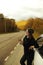 Stylish girl in swamp hat and black hoodie drinking coffee in craft glass on the side of an autumn forest road, traveling by car,