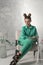 Stylish girl in turquoise shirt and trousers sits elegantly on chair in studio. Model with bun hairstyle and evening makeup, gray