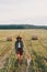 Stylish girl walking with her dog at hay bale in summer field in sunset. Young woman in hat traveling with pet in haystack field.