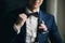 Stylish groom in blue suit with boutonniere with pink rose holding bow tie and posing near window in hotel room. Morning