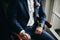 Stylish groom in blue suit, with bow tie and boutonniere with pink rose sitting in chair near window in hotel room. Morning