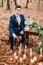 Stylish groom sits at the vintage table with cake and candles in autumn forest