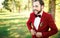 Stylish groom in tuxedo laughs suit marsala red, burgundy bow tie. Man buttoning his jacket outdoors. Professional