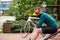 Stylish handsome guy cyclist rests on a wooden bench near bicycle in summer park.