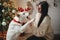 Stylish happy woman in cozy sweater playing with adorable dog under christmas tree with gifts and lights. Cute white dog giving