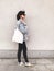 Stylish hipster girl in denim outfit and tote bag posing in city street on background of wall. Phone photo. Fashionable woman with