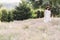 Stylish hipster girl in linen dress and hat relaxing in lavender field near tree, focus on lavender. Bohemian woman enjoying