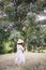 Stylish hipster girl in linen dress and hat relaxing in lavender field near tree. Happy bohemian woman enjoying summer vacation in