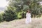 Stylish hipster girl in linen dress and hat relaxing in lavender field near tree. Happy bohemian woman enjoying summer vacation in