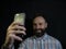 Stylish man with a beard and mustache smiles and takes a selfie on the phone on a black background