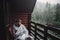 stylish man traveler in blanket relaxing on porch of wooden cabin in rainy day on background of woods in mountains. stylish