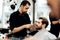 Stylish mustachioed barber dressed in a black shirt with a red bow tie scissors the hair of a young man in a barbershop