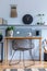 Stylish scandinavian living room interior with wooden desk, chair, wood panleing with shelf, table lamp, plants, black clock.