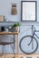 Stylish scandinavian living room with mokc up poster frame on the shelf, wooden desk, bicycle, office supplies.