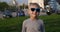 Stylish smiling boy blond in sunglasses stands on the lawn. The young schoolboy enjoys the holidays. City in the