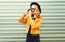 Stylish smiling woman with retro camera taking picture wearing yellow jacket, black round hat on metal wall