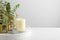 Stylish tender composition with burning candle and plants on white wooden table against light background, space for text