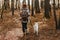 Stylish woman with backpack hiking with white dog in sunny autumn woods. Cute swiss shepherd puppy