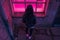 Stylish woman in black leather jacket looking at pink window on urban street, back view at night