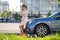 Stylish woman driver standing near her vehicle talking on cellphone on city street in summer