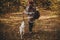 Stylish woman in hat and with backpack walking with cute dog in sunny autumn woods. Young female traveler hiking with swiss