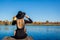 Stylish woman in hat sitting on river pier holding yellow leaves. Autumn landscape