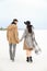 Stylish woman and man walking on snow and holding hands, wearing coat, grey scarf and hat.