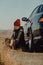 Stylish woman no face in warm gray coat and knitted Burgundy hat is sitting on ground near her car. Local auto-travel, life style