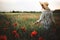 Stylish woman in rustic linen dress walking in summer meadow among poppy and wildflowers in sunset light. Atmospheric authentic