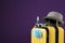 Stylish yellow suitcase with protective mask, sunglasses, hat and antiseptic spray on purple background, space for text.