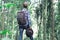 Stylish young man in plaid shirt with trendy leather backpack and felt hat in hand, standing in green forest. Escape into the wild