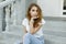 Stylish young pretty woman in a fashionable white t-shirt in trendy blue jeans sits on the vintage steps of the hotel outdoors.