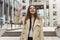 Stylish young smiling businesswoman wearing smart outfit stands in the middle of the street looking gorgeous waving her friend,