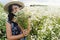 Stylish young woman in blue vintage dress and hat gathering white wildflowers in summer meadow. Beautiful happy girl holding white