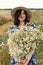 Stylish young woman in blue vintage dress and hat walking with white wildflowers in summer meadow. Happy beautiful girl smiling