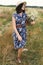 Stylish young woman in blue vintage dress and hat walking with white wildflowers in summer meadow. Happy beautiful girl smiling