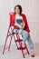 stylish young woman in white t shirt, jeans and red jacket and shoes on high heels is sitting on red ladder. studio