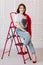 stylish young woman in white t shirt, jeans and red jacket and shoes on high heels is sitting on red ladder. studio