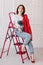 stylish young woman in white t shirt, jeans and red jacket and shoes on high heels is sitting on red ladder. studio