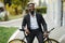 Stylishly dressed young African man smiling with earbeans lister voice from his cellphone while standing with his bicycle on a