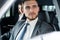 Success in motion. Handsome young man in full suit smiling while driving a car.