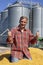 Successful Farmer Sitting in Trailer Full of Corn Seeds and Giving Thumbs Up in front of Farm Grain Bins