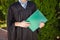 Successful graduate guy, in academic dresses, on graduation posing, holding a green folder and looking at it, farewell study, can