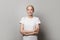 Successful optimistic young woman in white empty t-shirt standing with crossed arms on white studio wall background