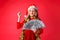 Successful teenage Girl in Santa hat and tinsel around her neck holding dollars in her hands and showing success gesture, on red