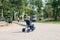 Summer in the city. In a children`s park with huge poplars along the path, a little blond boy rides a blue tricycle