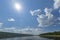 Summer day landscape with river, forest, clouds on the blue sky and sun.