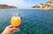 Summer drink cocktail of fresh orange juice with ice in the woman hand on the seascape background of Kolona beach Kythnos island C